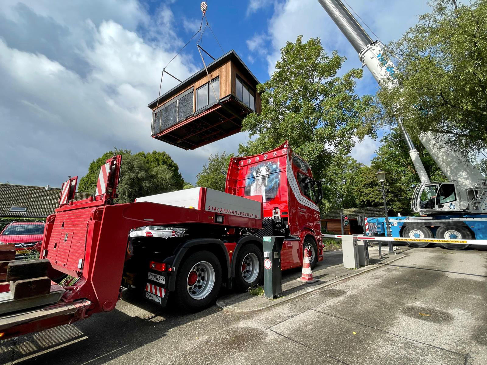 Een houten woning wordt met een kraan van de rode dieplader getild en op zijn plek gezet