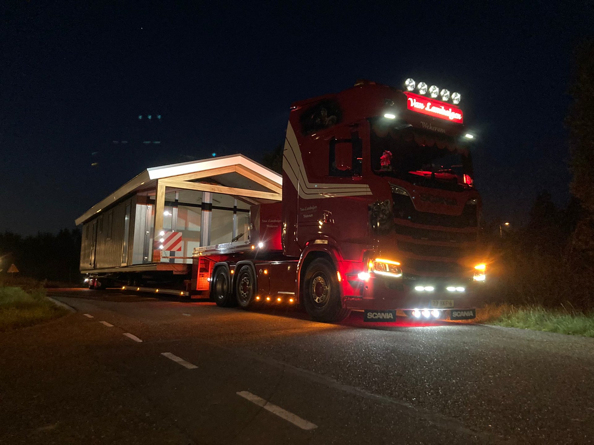 Een rode vrachtwagen met een brede woning op de dieplader, verlicht in het donker