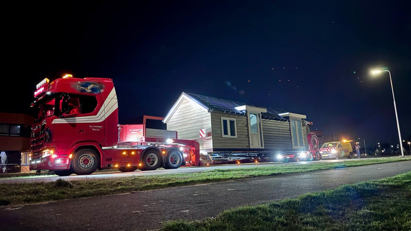 Een rode vrachtwagen vervoert 's avonds een woning, met een begeleidingsauto erachter
